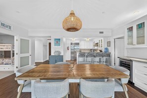 A spacious dining area with a wooden table and grey upholstered chairs, a hanging woven light fixture, and a kitchen with white cabinets and a built-in oven. - Residence 109 at The Grand (Wildwood)