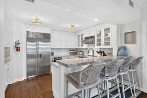 A modern kitchen with white cabinetry, stainless steel appliances, and a bar area with wicker chairs. - Residence 109 at The Grand (Wildwood)
