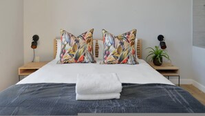 Ein Bett mit weißer Decke, zwei Kissen mit buntem Blattmuster, ein gefaltetes weißes Handtuch, ein Nachttisch mit Lampe und eine Topfpflanze auf dem Nachtkästchen.
