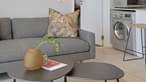 Ein modernes Wohnzimmer mit einem grauen Sofa, einem kleinen runden Tisch, einer dekorativen Vase und einer Waschmaschine im Hintergrund.