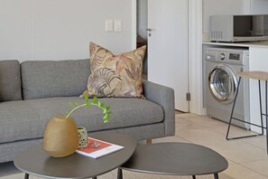 Ein modernes Wohnzimmer mit einem grauen Sofa, einem kleinen runden Tisch, einer dekorativen Vase und einer Waschmaschine im Hintergrund.