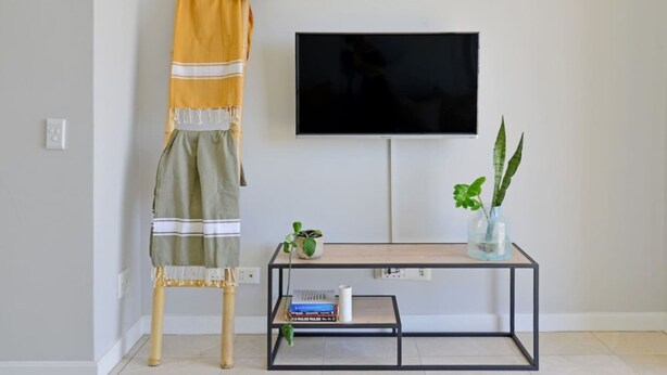 Ein Zimmer mit einem an der Wand befestigten Fernseher, einem kleinen Tisch mit Pflanze und Büchern sowie einem Beistelltisch mit Hut und Handtuch.