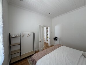 Une chambre avec un lit, une échelle en bois, un ventilateur et une porte donnant sur un couloir avec une porte blanche et des étagères.
