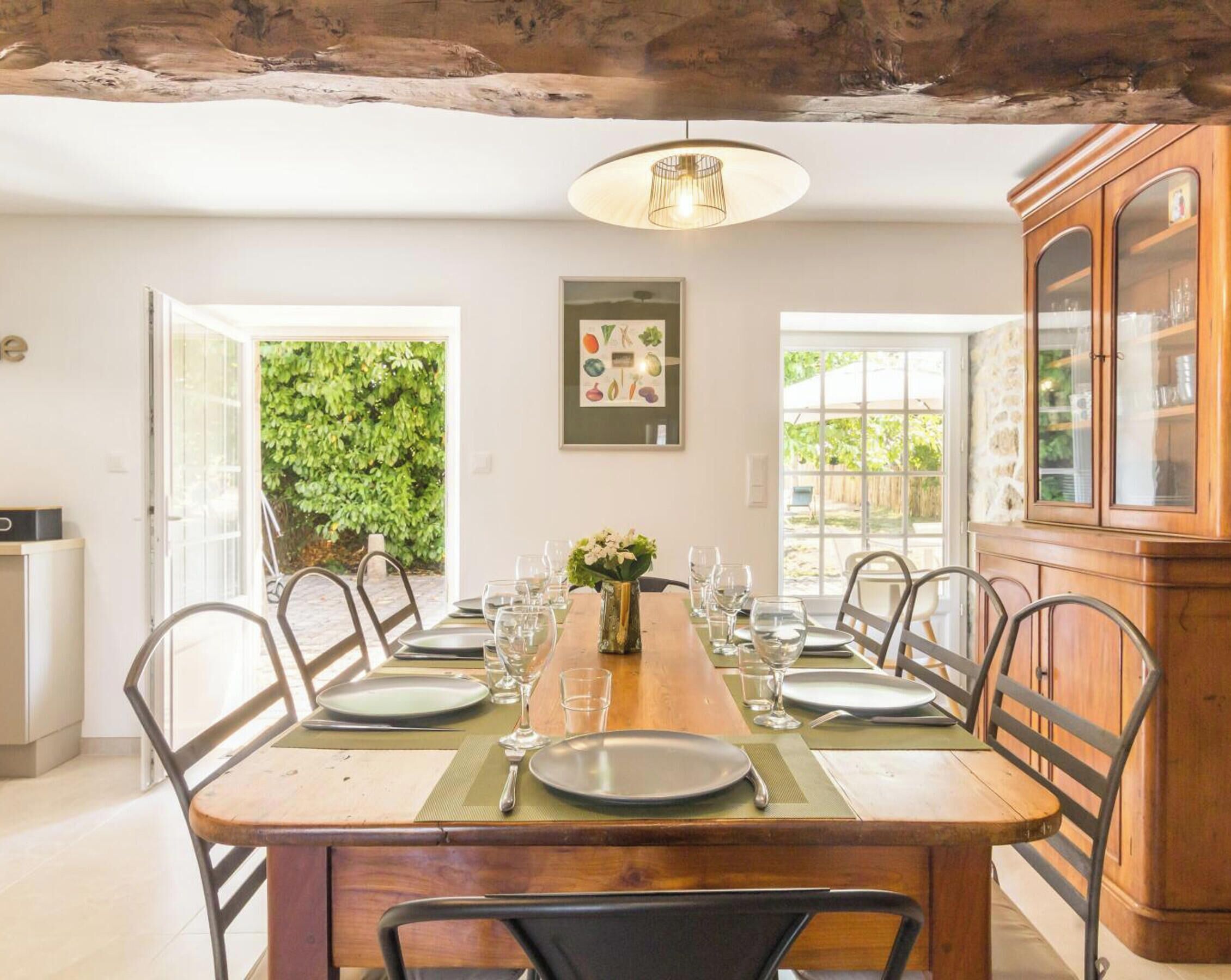 A dining area with a wooden table set for six, surrounded by metal chairs, with a view of greenery outside.