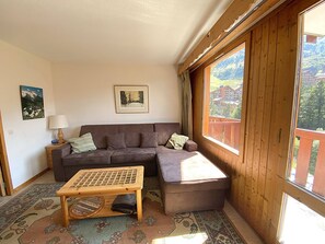 Ein gemütliches Zimmer mit einem braunen Sofa, einem kleinen Holztisch und einem Fenster mit Blick auf Gebäude.