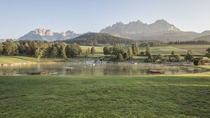 Blick auf einen See mit einer grünen Vordergrundwiese, einem kleinen Steg und einem Bergpanorama im Hintergrund.