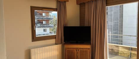A room with a television on a wooden stand, a window with curtains, a radiator, and a view of a building outside.