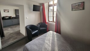 A room with a bed, two chairs, a TV, a window with curtains, a kitchen area, and a painting on the wall. - Charming apartment in city center - 2 guests (Luxeuil-les-Bains)