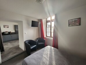 Ein Zimmer mit einem Bett, zwei Stühlen, einem Fernseher, einem Fenster mit Vorhängen, einer Küchenzeile und einem Bild an der Wand.