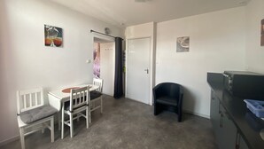 A compact room with a dining table and chairs, a black chair, a kitchenette, and a door. - Charming apartment in city center - 2 guests (Luxeuil-les-Bains)