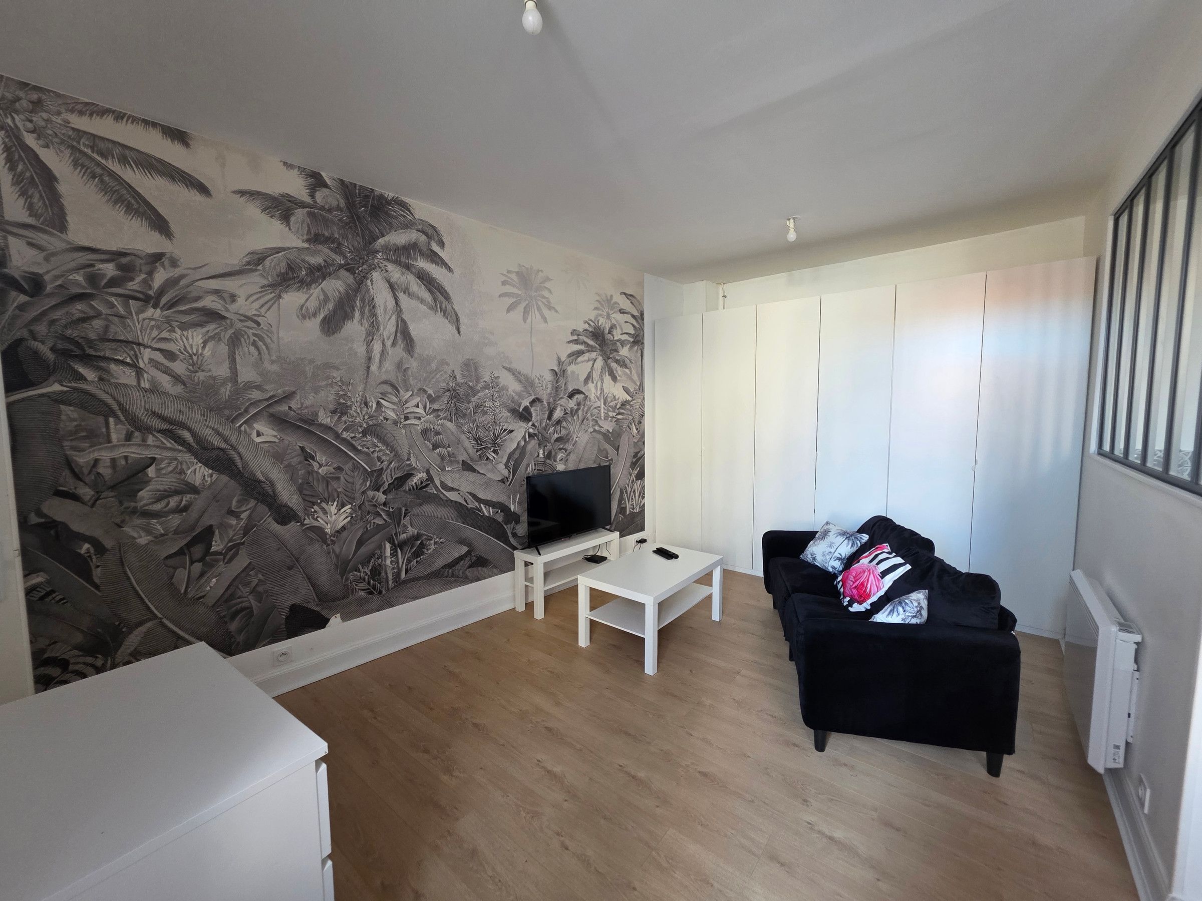 Una sala de estar con un televisor sobre un soporte blanco, un sofá negro con un cojín estampado y un mural de follaje tropical.