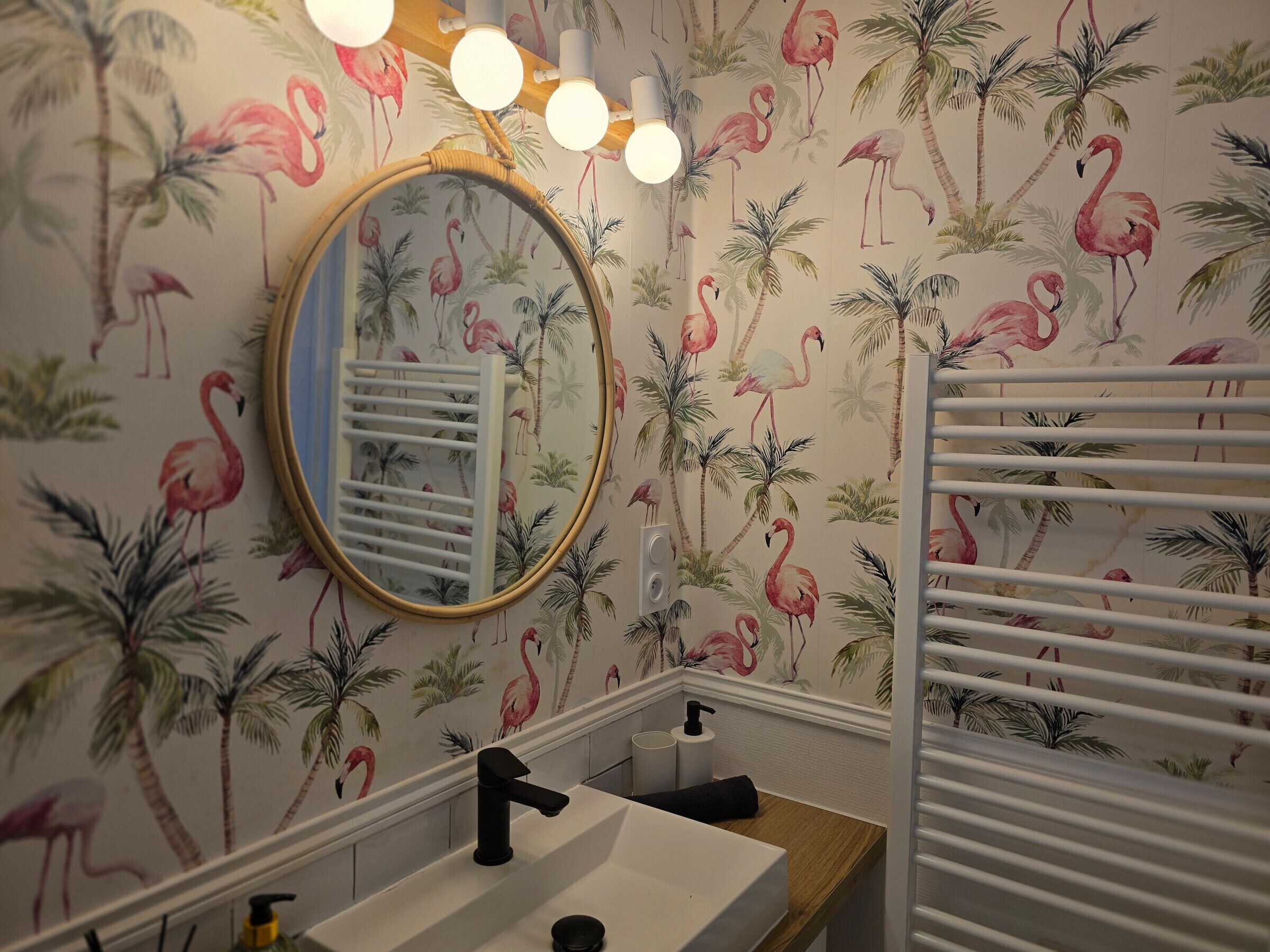Un baño con lavamanos, espejo y toallero frente a una pared con papel tapiz floral que muestra flamencos y palmeras.
