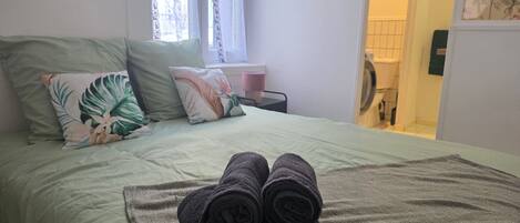 Una cama bien hecha con una colcha verde y dos almohadas, una toalla enrollada sobre la cama y una ventana con cortinas traslúcidas.