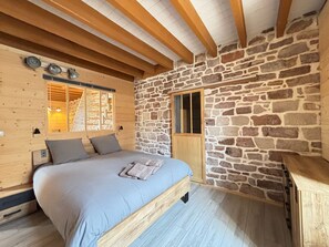 A bedroom with a stone wall, wooden ceiling beams, a bed with grey bedding, and a window with a view. - Charming apartment in Luxeuil-les-Bains (Luxeuil-les-Bains)