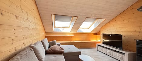 A loft-style room with a sofa, ottoman, TV, and wooden beams.