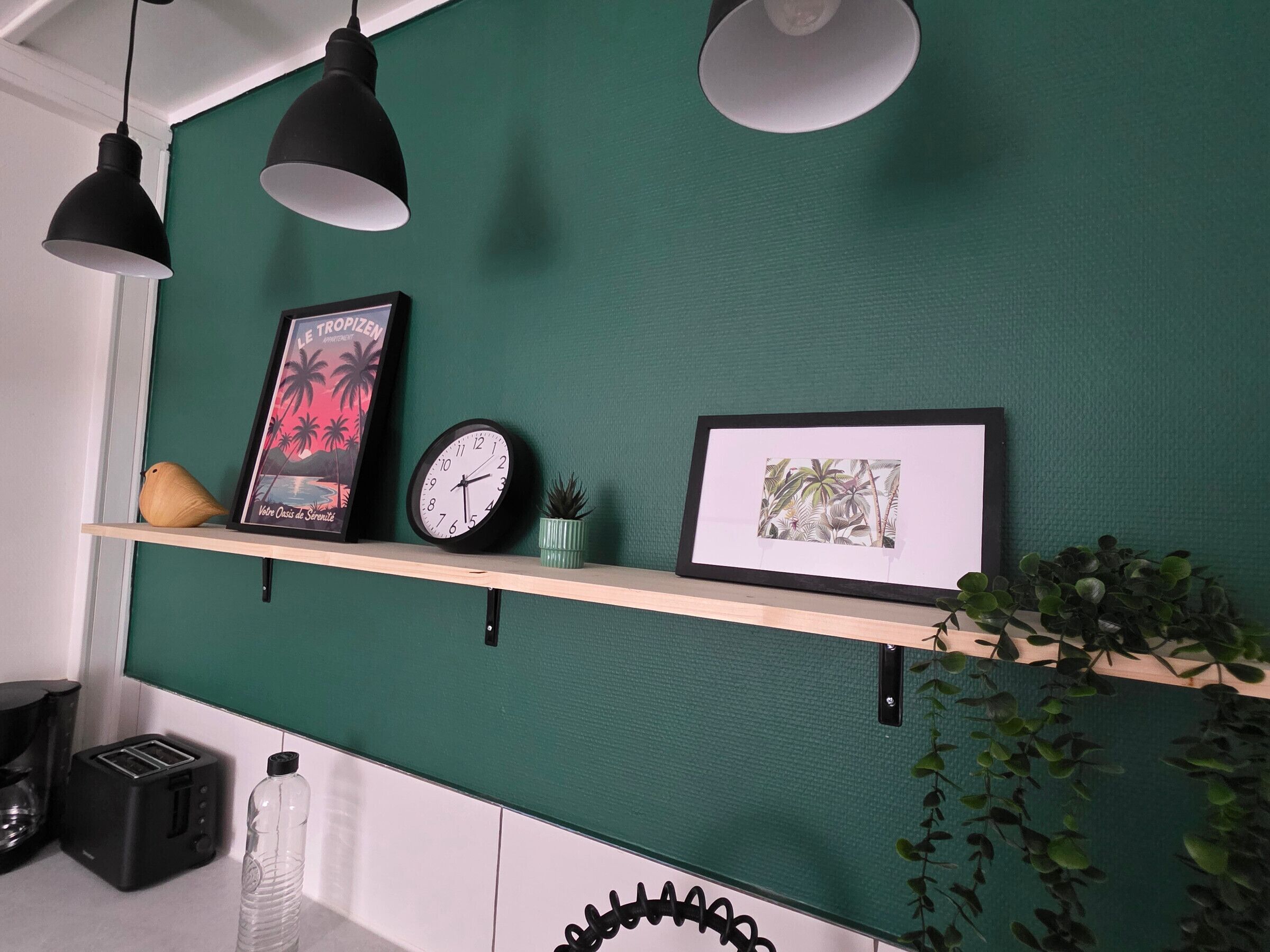 Una cocina con pared verde, estante con reloj, cuadro enmarco y planta en maceta.