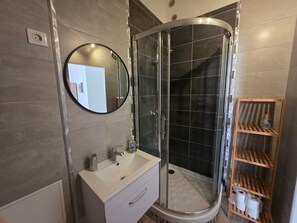 Ein modernes Badezimmer mit einer in Glas abgeschirmten Dusche, einem weißen Waschbecken, einem Spiegel und einem Holzregal mit Pflegeprodukten.