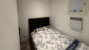 Ein kleines Zimmer mit einem Einzelbett, einer schlichten weißen Wand, einem Fenster mit Jalousien und einer Wand-Klimaanlage.