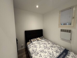 Ein kleines Zimmer mit einem Einzelbett, einer schlichten weißen Wand, einem Fenster mit Jalousien und einer Wand-Klimaanlage.