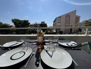 Une table dressée pour deux personnes, avec des assiettes portant la lettre « V », une bouteille de vin et des verres, le tout sur un balcon donnant sur des immeubles.