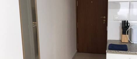 A narrow hallway with a wooden door at the end, a white cabinet with a towel on top, and a small kitchen area with a sink and utensils.