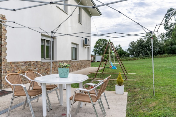 An outdoor patio area with a round table and four wicker chairs, a potted plant, and a swing set.