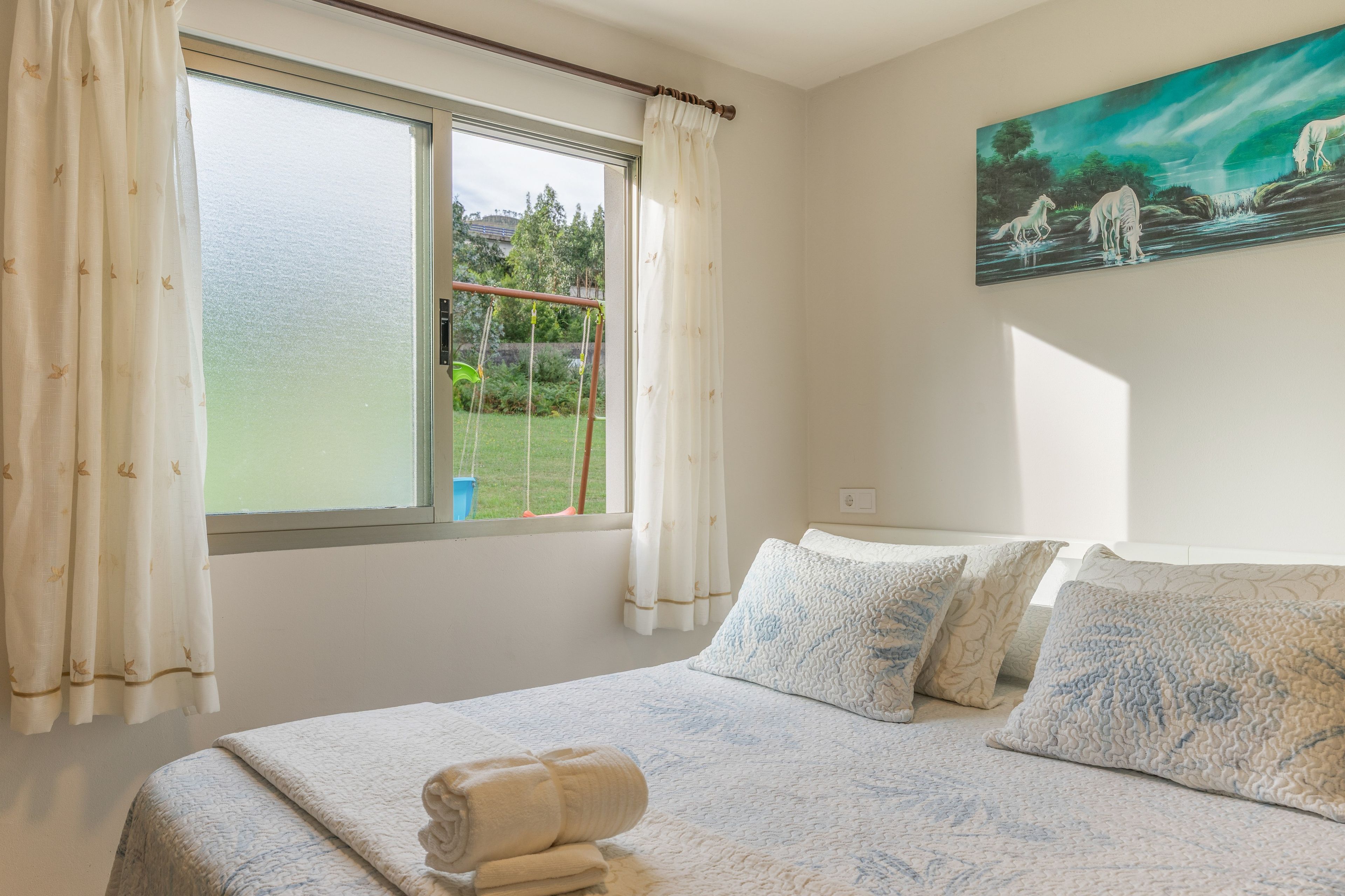Un dormitorio con cama, almohadas, un cuadro y una ventana con cortinas.
