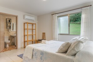 A bedroom with a bed, a wooden ladder, a window with curtains, a bathroom with a toilet, and an air conditioning unit.