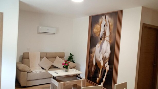 A living room with a beige sofa, a small table with a vase of flowers, and a large painting of a horse on the wall.