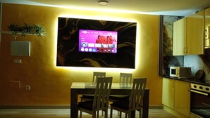 A modern dining area with a flat-screen TV mounted on the wall, a dining table with chairs, and a kitchen area with cabinets and appliances.