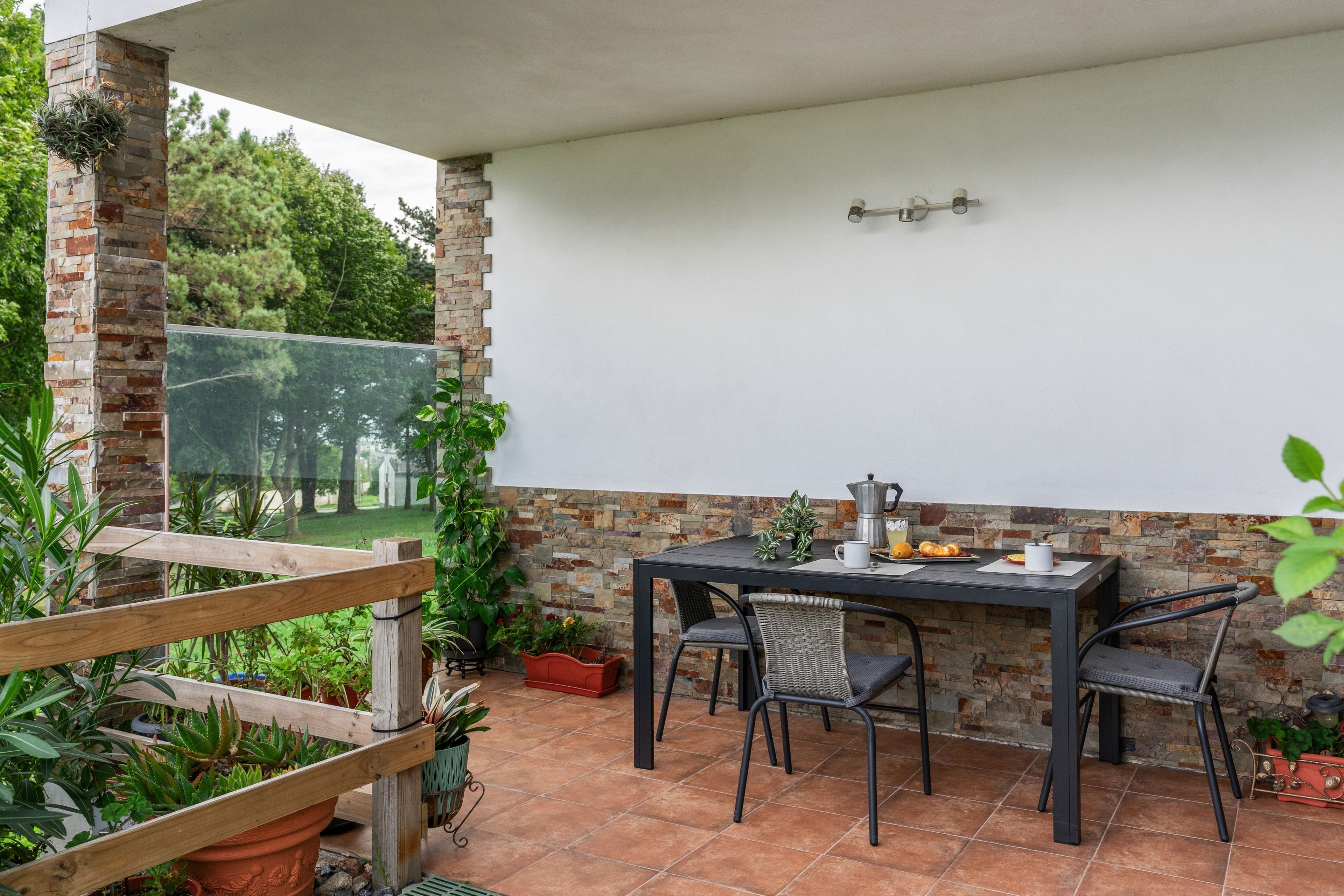 Een terras buiten met een tafel en stoelen, omgeven door potplanten en een stenen muur.