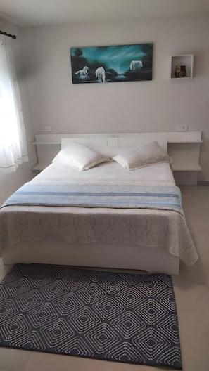 A bedroom with a bed, pillows, a blanket, a painting, a shelf, and a rug.