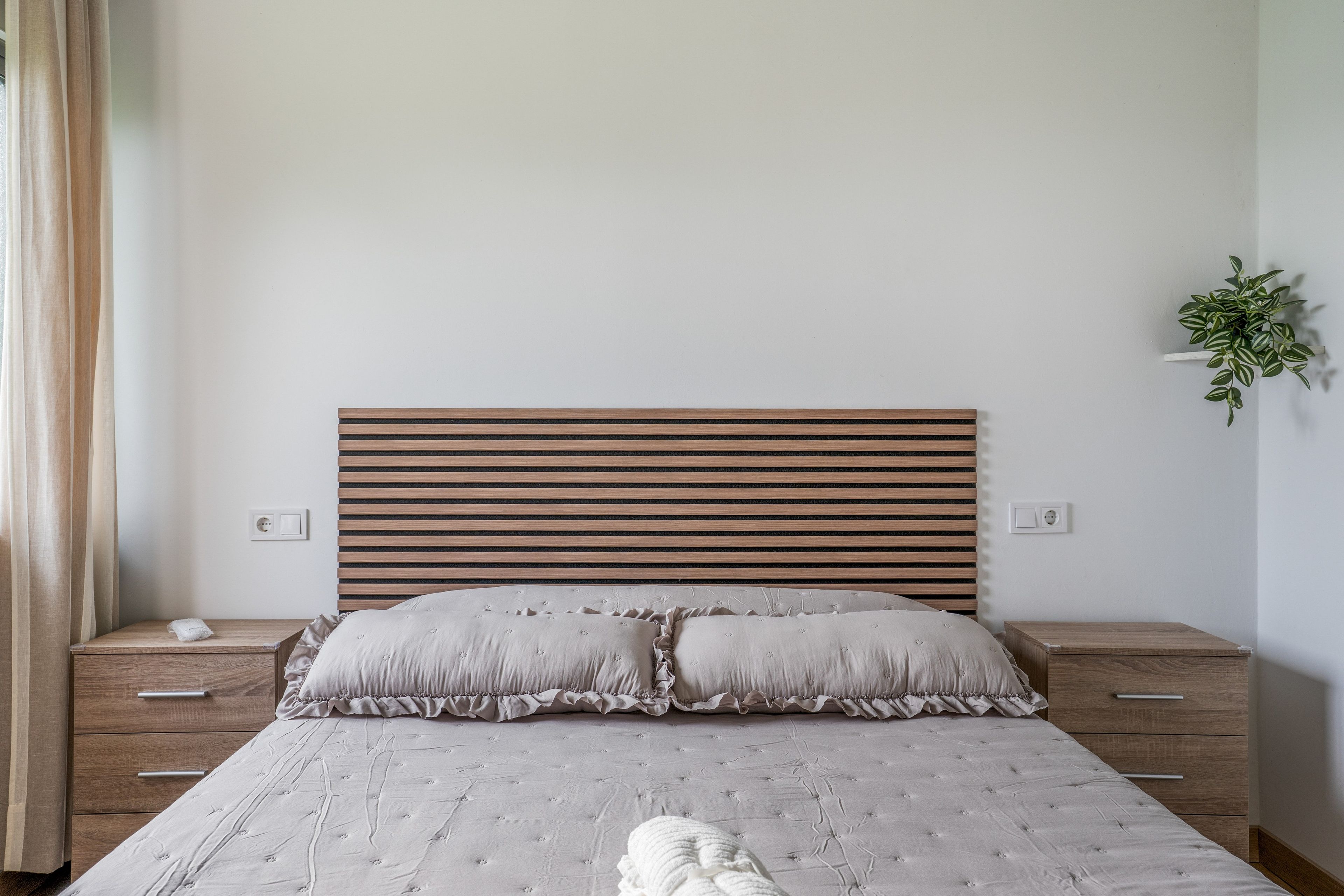 Een slaapkamer met een houten lattenkap, een bed met een witte dekbedovertrek en geplooide kussens, twee nachtkastjes met lades en een wandplant.