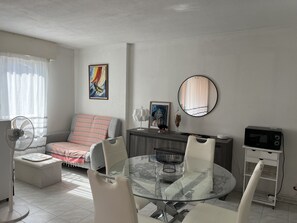 Ein modernes Wohnzimmer mit Glastisch, weißen Stühlen, einer Couch mit gestreiften Kissen, einem Standventilator, einem gerahmten Gemälde, einem dekorativen Spiegel und einer Mikrowelle.