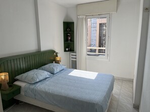 Een slaapkamer met een bed, nachtlampen, een groen rek met decoratieve objecten, een raam met uitzicht op gebouwen, en een wit gordijn.