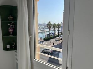 Blick aus einem Fenster auf eine grüne Ablage mit Dekoration, einen klaren Himmel und einen Yachthafen mit Booten und Menschen.