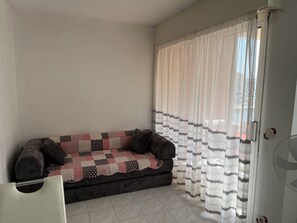 Ein Zimmer mit einer Couch, die mit einer gemusterten Decke bedeckt ist, einem weißen Stuhl und einem Fenster mit transparenten Vorhängen.