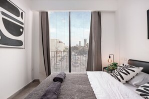 Habitación de hotel moderna con una cama grande, vistas de la ciudad a través de ventanales del suelo al techo, una mesita auxiliar con una planta y un aplique en la pared.
