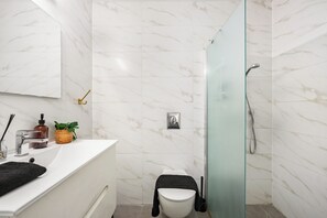 Un baño moderno con pared de mármol blanco, cabina de ducha de vidrio, lavamanos blanco y una toalla negra, además de una pequeña planta en la encimera.
