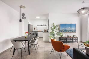 Una cocina moderna con armarios blancos, una zona de comedor con una mesa oscura y sillas blancas, y una sala de estar con un sofá y un televisor.