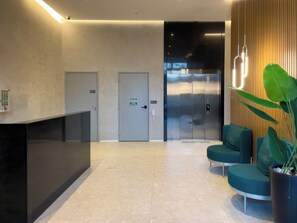 Zona de recepción con mostrador, dos puertas, un ascensor moderno y un área de estar con planta en maceta.