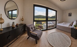 Un salon moderne comprenant un canapé, un fauteuil, une grande fenêtre donnant sur un paysage verdoyant, un tapis rond et une console noire agrémentée d’objets décoratifs.