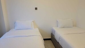 Zwei Einzelbetten mit weißer Bettwäsche in einem Raum mit schlichten Wänden und einer kleinen Lampe.