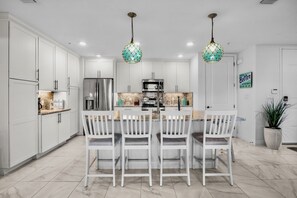 A modern kitchen with white cabinetry, a central island with bar stools, stainless steel appliances, and decorative hanging lights. - Sister’s Paradise by AMI Locals (Bradenton)