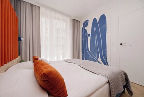 Une chambre d’hôtel avec un lit, une grande fenêtre avec des rideaux légers, un mur orné d’un motif abstrait bleu et une porte.