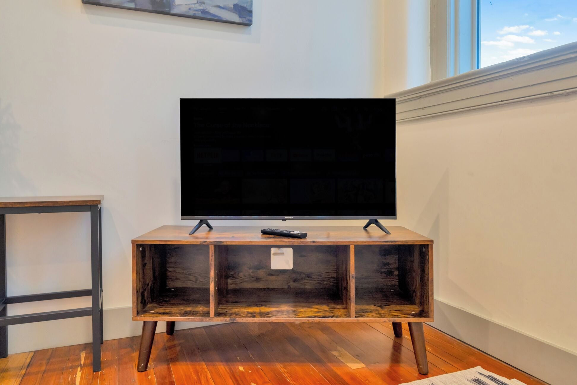 A television on a wooden stand with a remote control on top, positioned against a white wall with a window.
