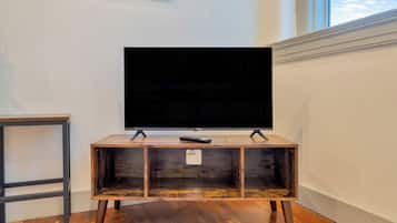 A television on a wooden stand with a remote control on top, positioned against a white wall with a window.