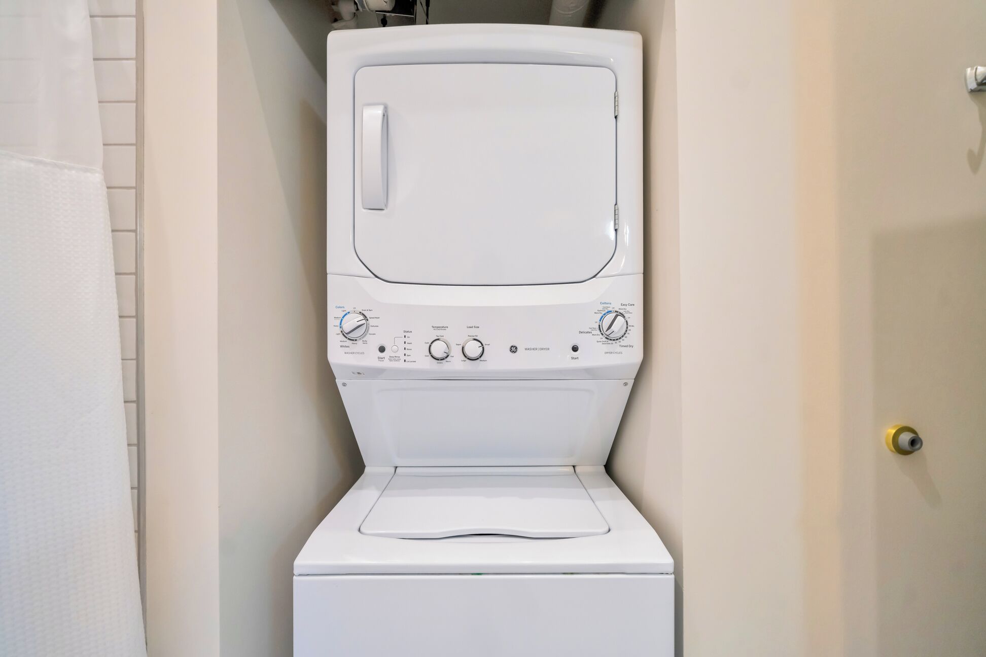 Une machine à laver à chargement frontal, de couleur blanche, se trouve dans une salle de bain dont un mur est recouvert de carreaux et qui possède une porte.