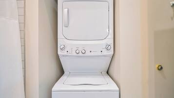 Une machine à laver à chargement frontal, de couleur blanche, se trouve dans une salle de bain dont un mur est recouvert de carreaux et qui possède une porte.