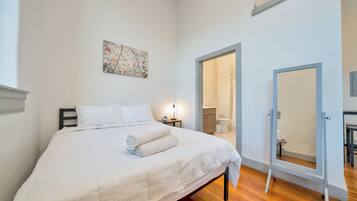 A bedroom with a bed, a bedside table, a nightstand, a mirror, and an open door leading to a bathroom.